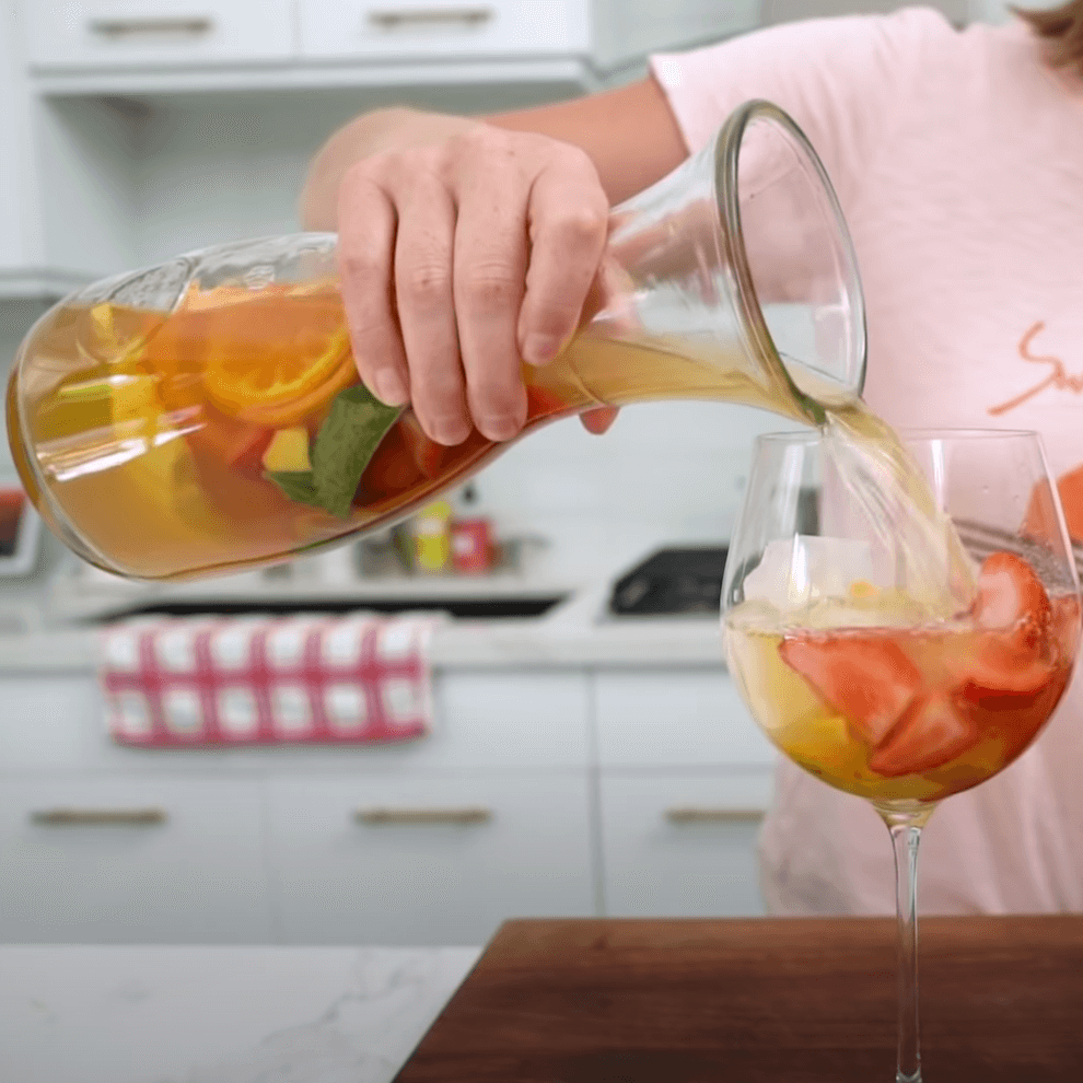 Hand pouring fruit-infused sangria into a wine glass in a kitchen setting.