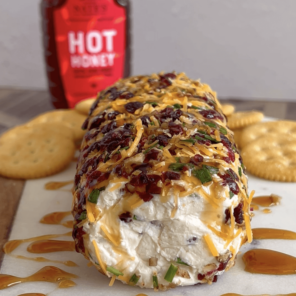 Cream cheese log dressed with pecans, cranberries, cheddar cheese and chives with crackers and Nate’s hot honey in background.