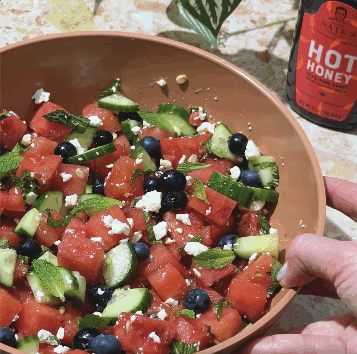 Bowl of watermelon salad with cucumber, blueberries, feta, and mint, drizzled with hot honey.
