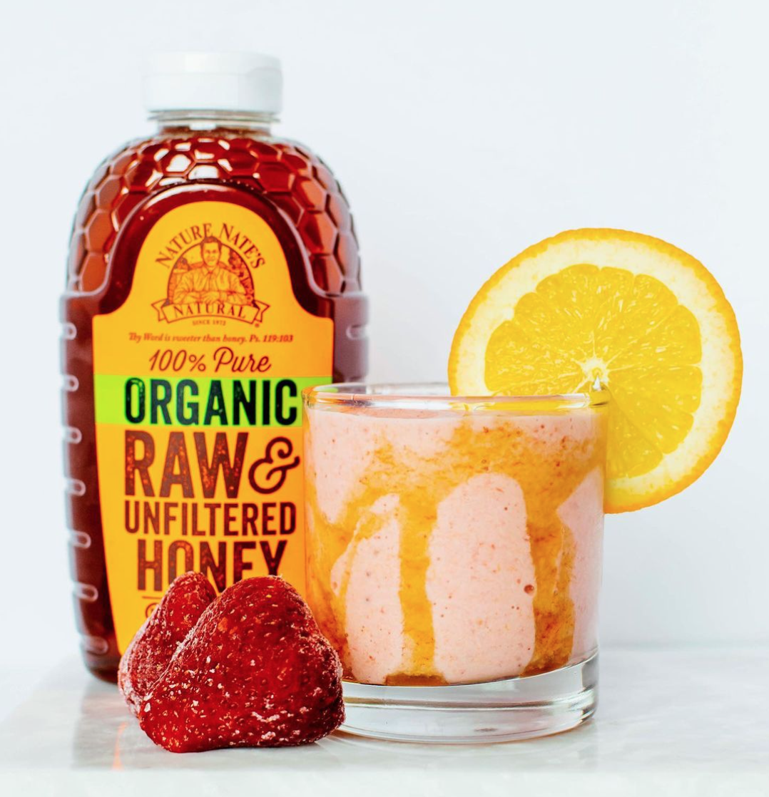 Bottle of Nate's 100% pure organic raw & unfiltered honey, and a glass with a pink smoothie and a strawberry garnish.