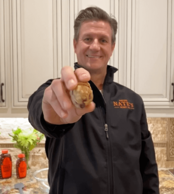 The man Nature Nate, smiling and offering a cookie, wearing a jacket with NATE'S logo, with bottles of Nate's honey.