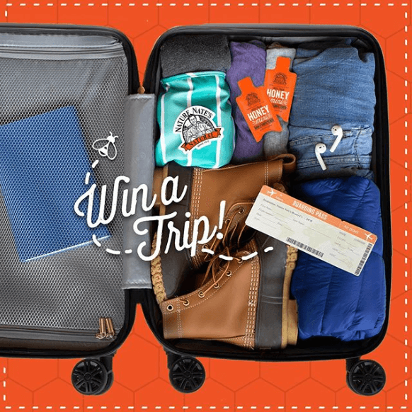An open suitcase with items for a trip: clothes, a ticket and a bottle of honey with the text Win a Trip!