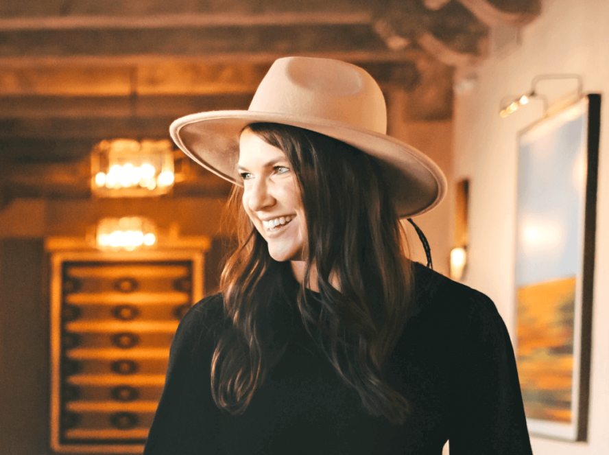 A smiling woman in a black outfit and beige hat, standing indoors with warm lighting and art in the background.