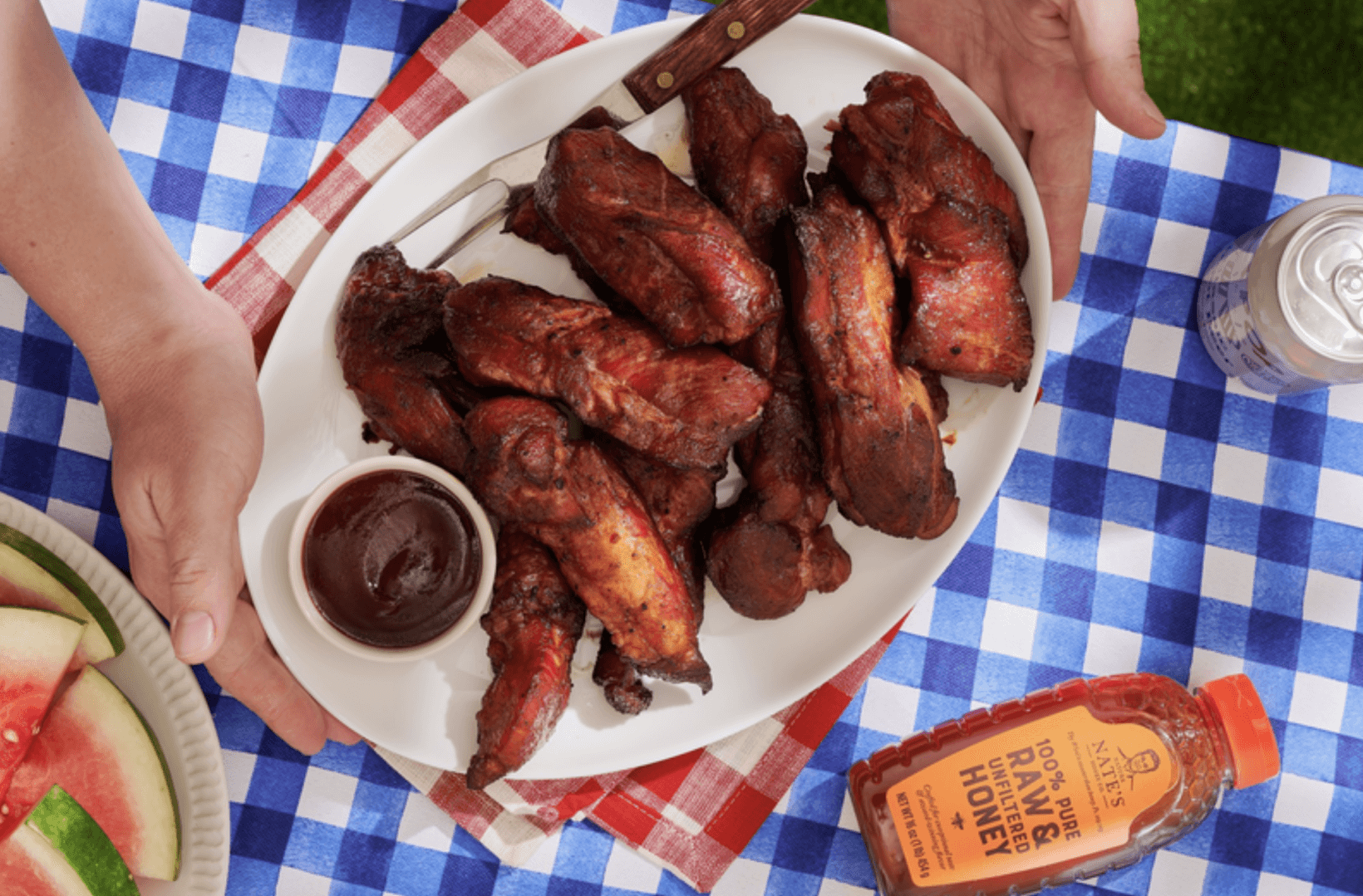 Hands serving a plate of BBQ pork ribs with a side of sauce and a Nate’s honey bottle, on a blue and white blanket.