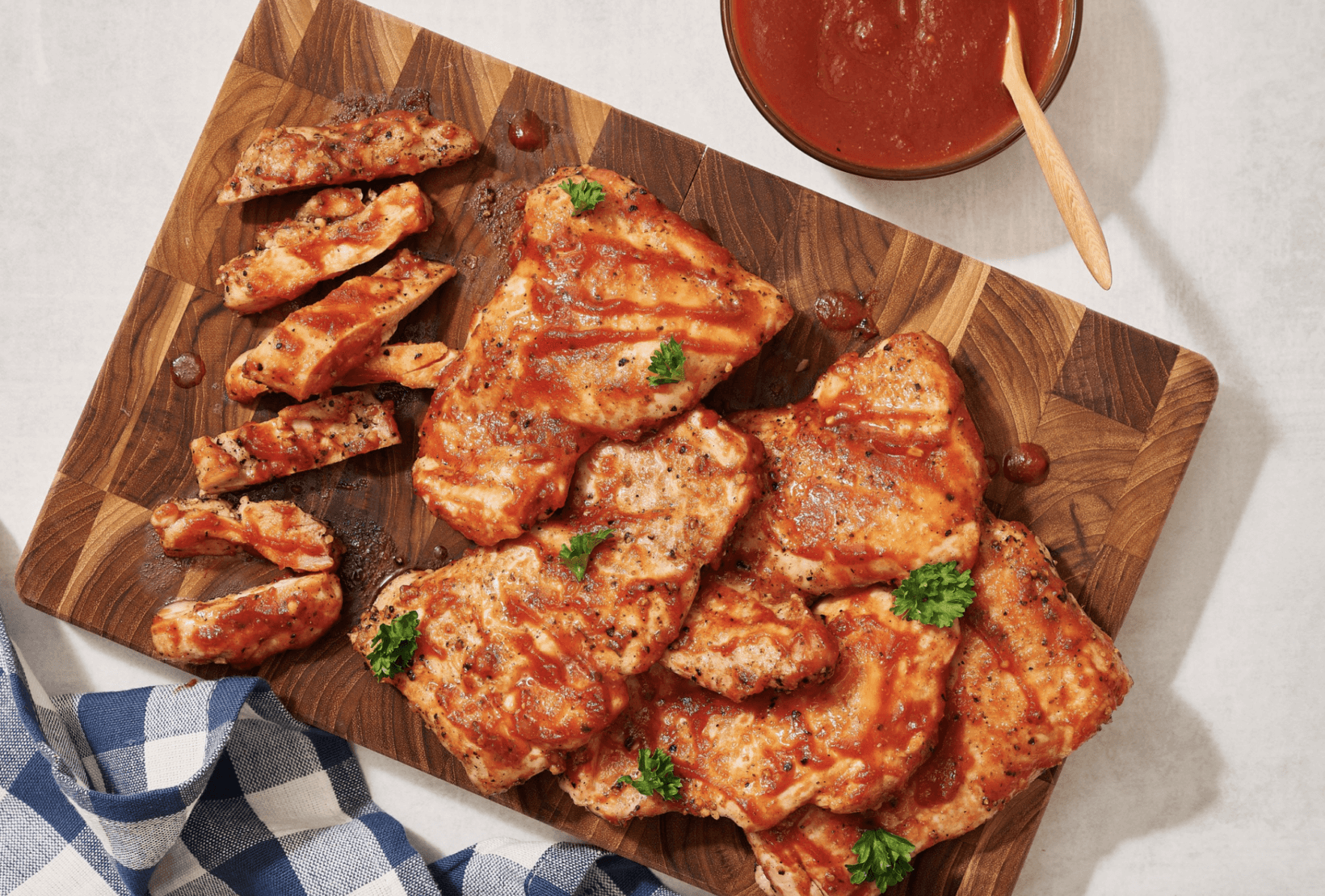 Grilled chicken glazed with honey barbeque sauce on a wooden board with a side of sauce, next to a checkered napkin.