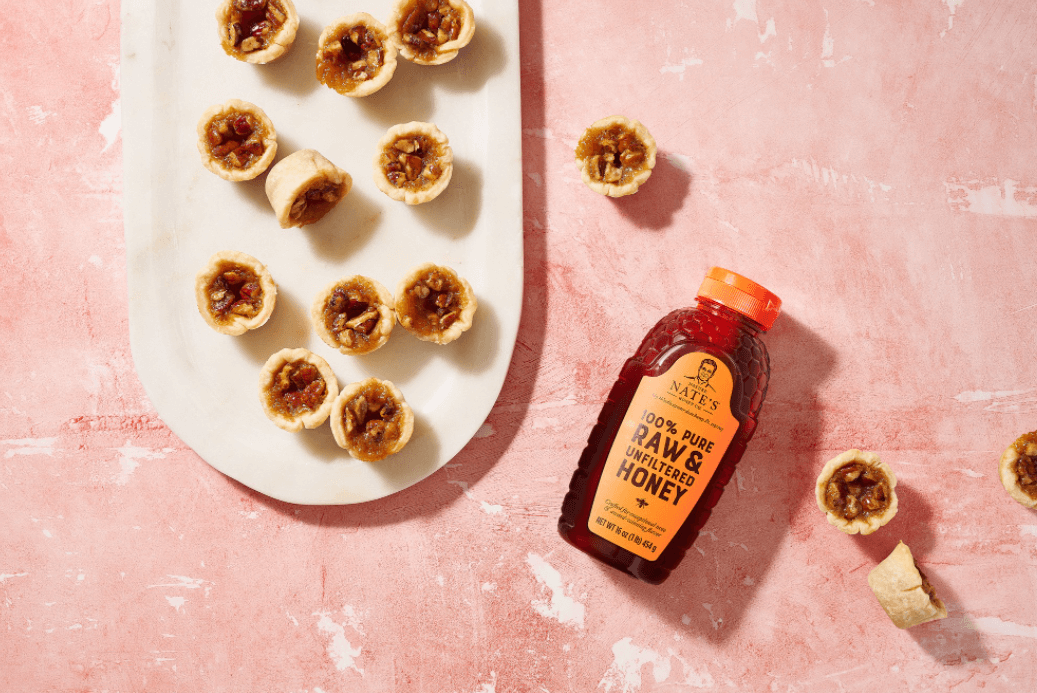 Mini pecan tarts on an oval plate and a bottle of Nate’s raw & unfiltered honey on a textured pink surface.