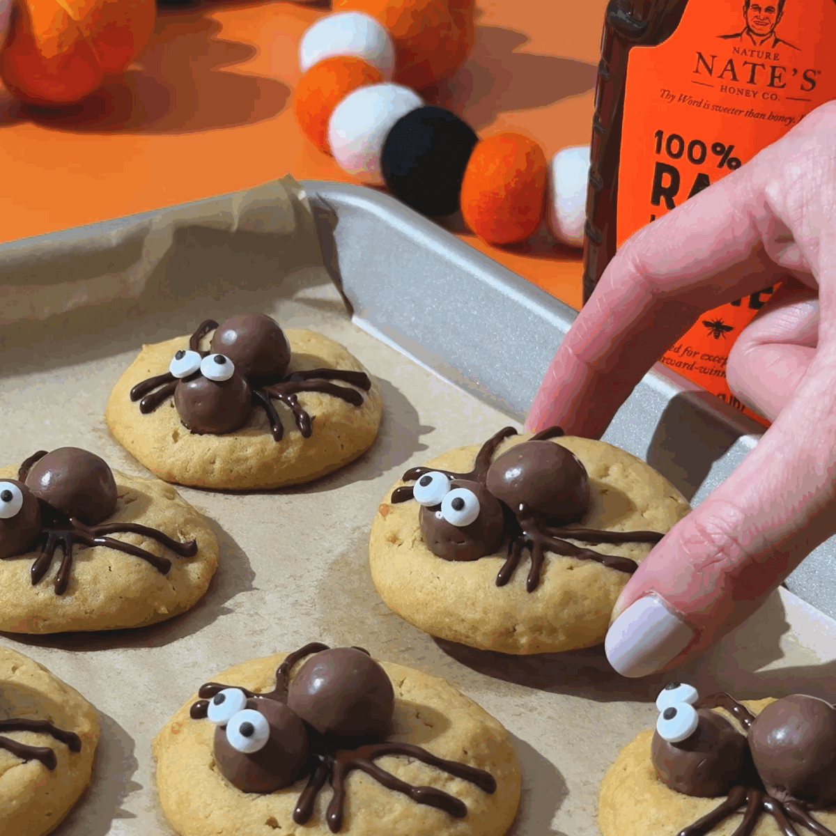 Spider cookies with chocolate bodies and candy eyes on a baking sheet, with a hand picking one up.