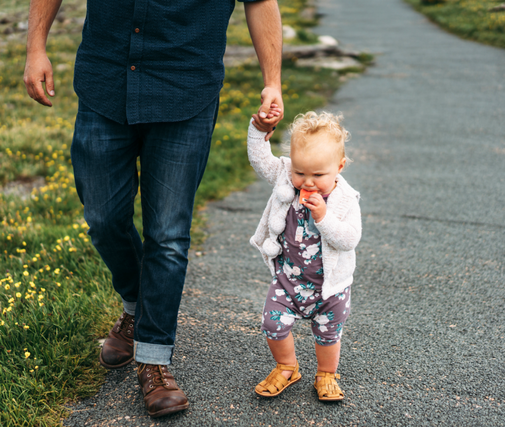 Toddler enjoying honey from a Nate's honey mini, holding adult's hand, outdoors on a path with grass.